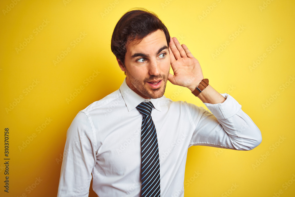 Young handsome businessman wearing elegant shirt and tie over isolated yellow background smiling with hand over ear listening an hearing to rumor or gossip. Deafness concept. Young handsome businessman wearing elegant shirt and tie over isolated yellow background smiling with hand over ear listening an hearing to rumor or gossip. Deafness concept.