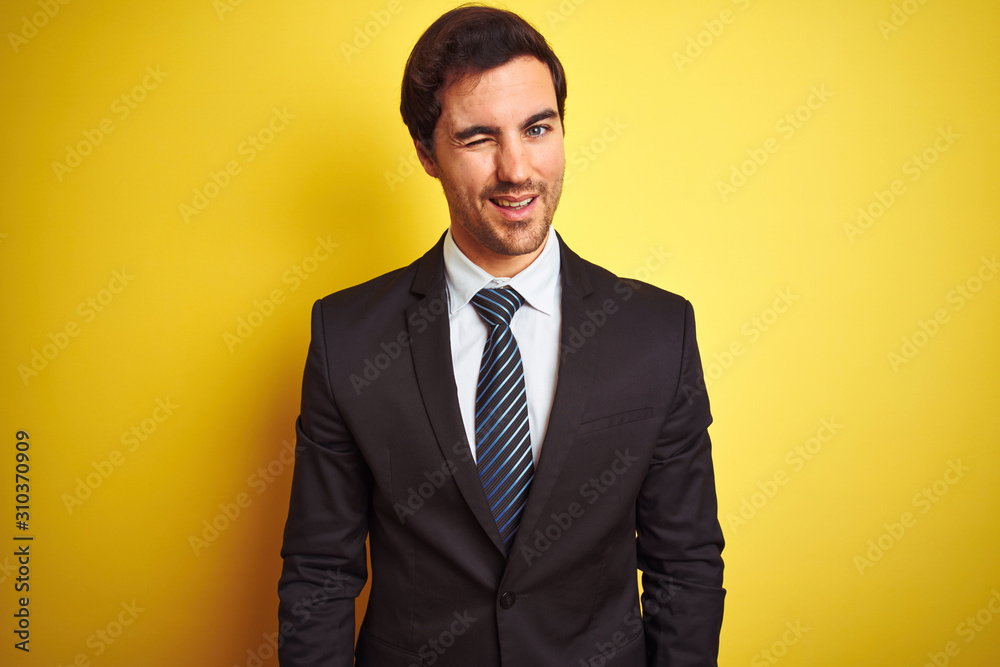 Young handsome businessman wearing suit and tie standing over isolated yellow background winking looking at the camera with sexy expression, cheerful and happy face. Young handsome businessman wearing suit and tie standing over isolated yellow background winking looking at the camera with sexy expression, cheerful and happy face.