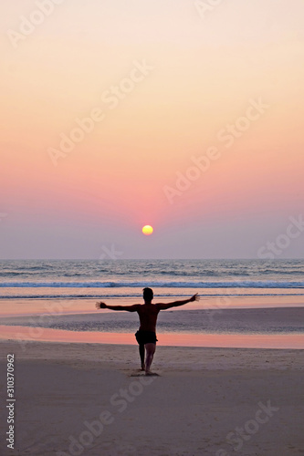 An unrecognizable man doing a yoga position sun salutation on an empty beach at sunset