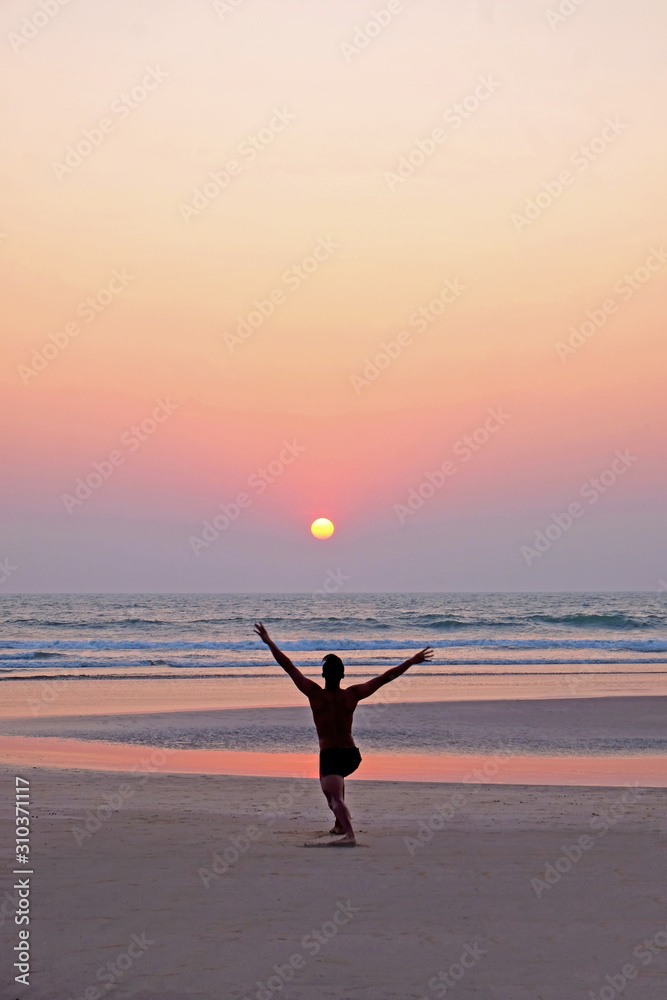 An unrecognizable man doing a yoga position sun salutation on an empty beach at sunset