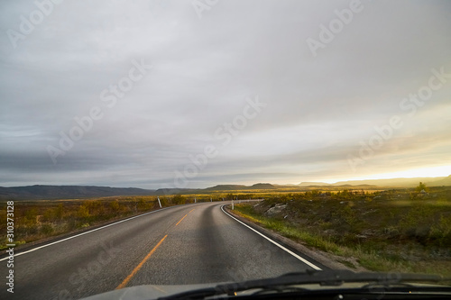 View on grey road, yellow surise and dramatic clouds from the car front window in early mourning. Driving car during sunshine in the tundra in Norway. Background of nature landscape and a road