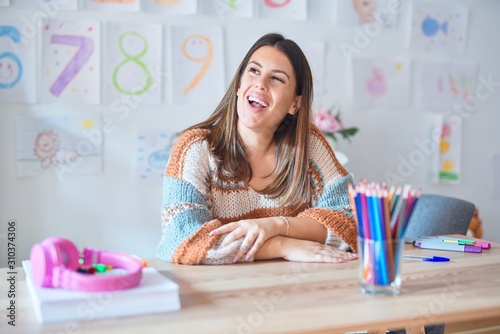 Young beautiful teacher woman wearing sweater and glasses sitting on desk at kindergarten looking away to side with smile on face, natural expression. Laughing confident.