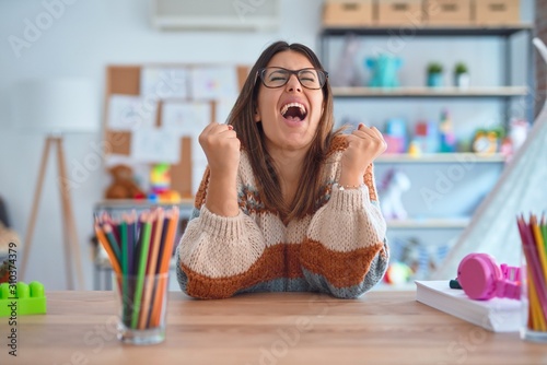 Young beautiful teacher woman wearing sweater and glasses sitting on desk at kindergarten very happy and excited doing winner gesture with arms raised, smiling and screaming for success. Celebration