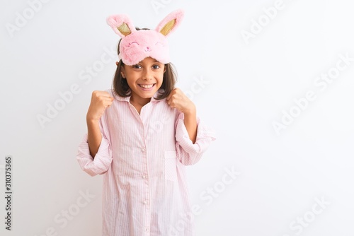 Beautiful child girl wearing sleep mask and pajama standing over isolated white background celebrating surprised and amazed for success with arms raised and open eyes. Winner concept.