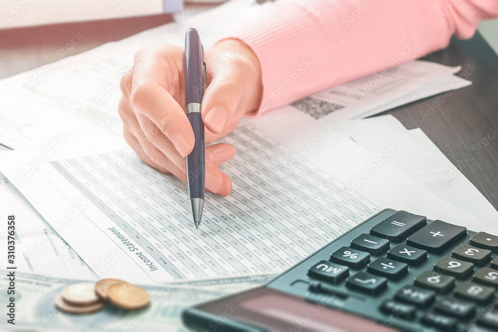 Closeup side view on the hand of female accountant who is holding a pen and working in an office with a stack of documents and tables. Calculation of taxes, profits and expenses, saving money concept Closeup side view on the hand of female accountant who is holding a pen and working in an office with a stack of documents and tables. Calculation of taxes, profits and expenses, saving money concept