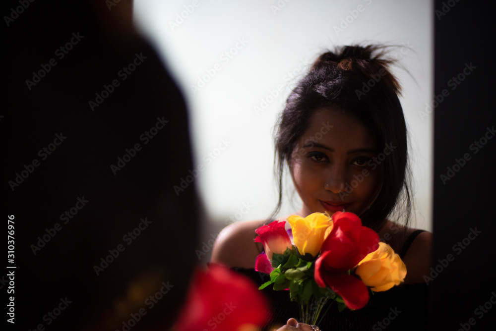 An young and attractive Indian  brunette woman in western wear standing in front of a mirror with a bunch of flowers. Indian lifestyle.