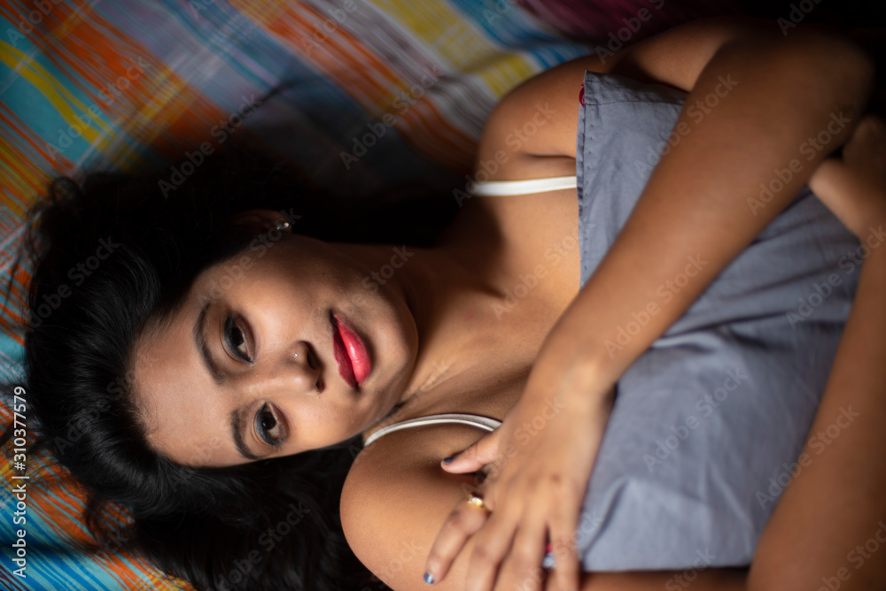 An young and attractive Indian Bengali brunette woman in white sleeping wear is lying on a bed with a blanket in her room in colorful background. Indian lifestyle. An young and attractive Indian Bengali brunette woman in white sleeping wear is lying on a bed with a blanket in her room in colorful background. Indian lifestyle.
