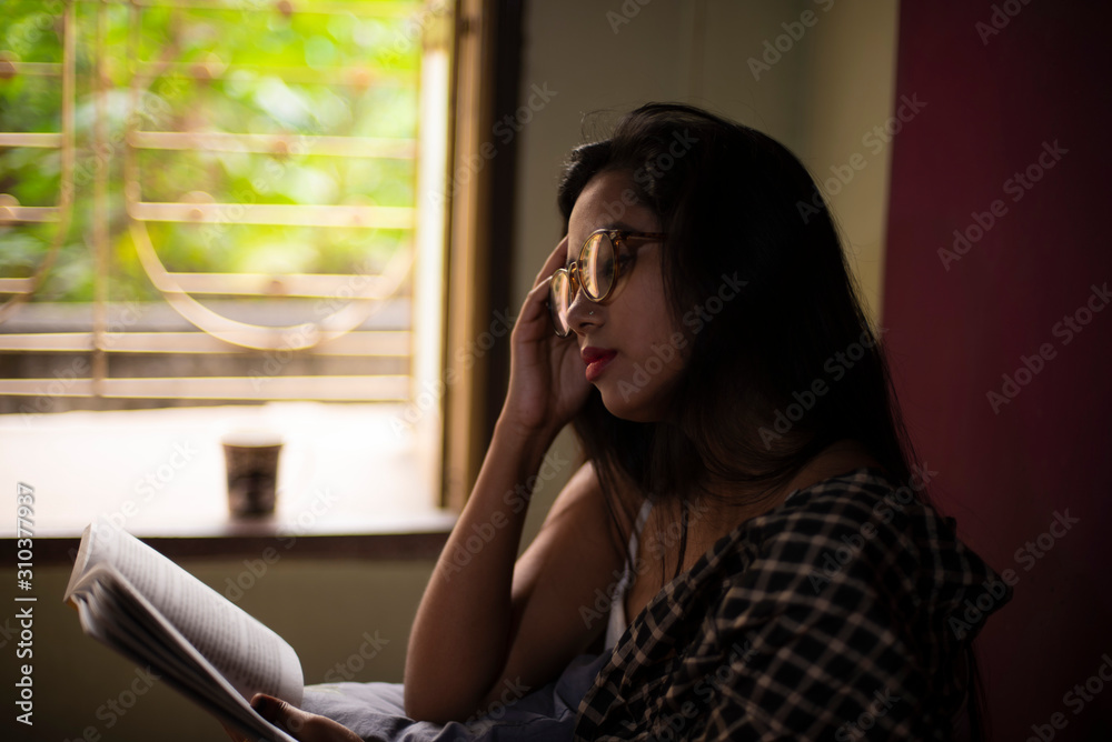 A young and attractive Indian Bengali brunette woman in white sleeping wear with a coffee/tea cup reading book on a bed in front of a window inside in her room. Indian lifestyle. A young and attractive Indian Bengali brunette woman in white sleeping wear with a coffee/tea cup reading book on a bed in front of a window inside in her room. Indian lifestyle.