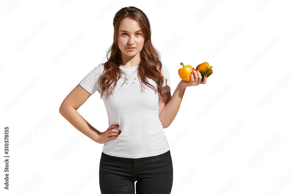 Veganism as a way of living is demonstrated by a pretty brunette girl in a white tee and black jeans. The healthy looking lady is showing two yellow and green peppers in her hand. Veganism as a way of living is demonstrated by a pretty brunette girl in a white tee and black jeans. The healthy looking lady is showing two yellow and green peppers in her hand.