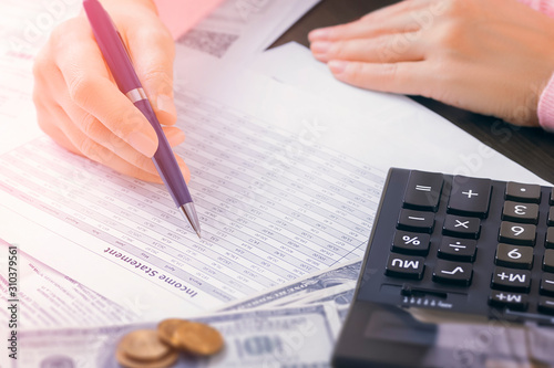 Closeup of female accountant hands in office who is holding a pen and is looking for data in a table. Steck of documents, calculator and dollars in workplace. Profit analysis, tax and payment concept