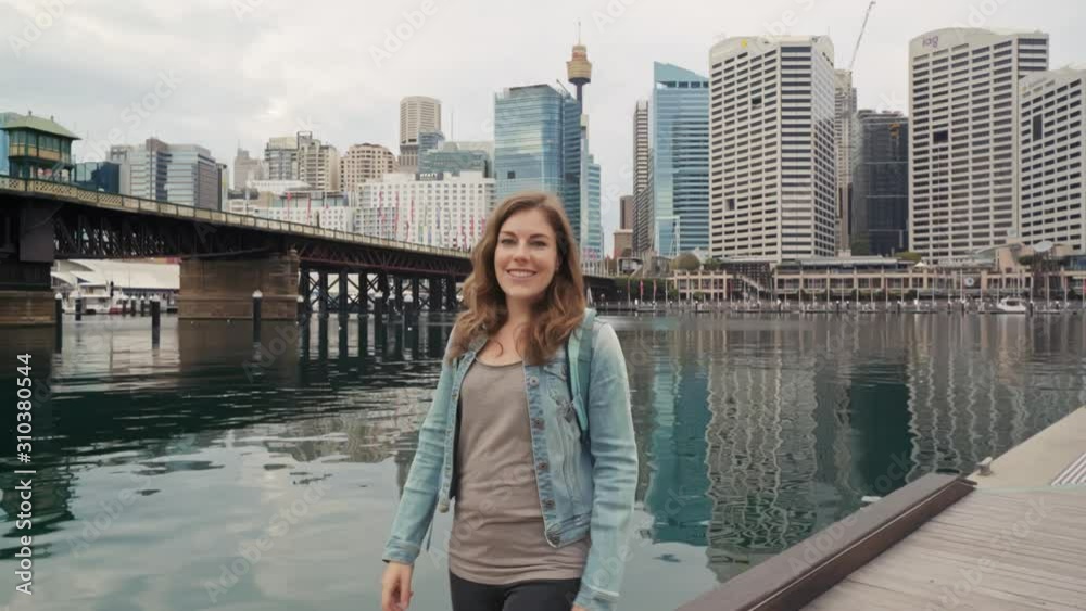 Smiling young woman walking down a pier flirtatiously looking at a camera with Sidney Australia skyline in the background Smiling young woman walking down a pier flirtatiously looking at a camera with Sidney Australia skyline in the background