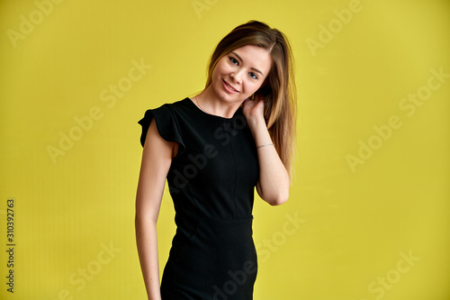 Portrait of a pretty young smiling girl on a yellow background in a black dress with long straight hair. Standing right in front of the camera, Shows emotions, talks in different poses.
