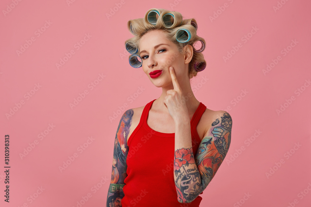 Close-up of pretty young blonde tattooed female with curlers on her head and red lips looking cunningly at camera and smiling slightly, dressed in casual clothes while standing over pink background
