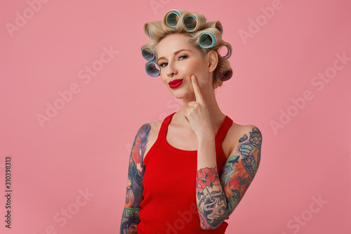 Close-up of pretty young blonde tattooed female with curlers on her head and red lips looking cunningly at camera and smiling slightly, dressed in casual clothes while standing over pink background