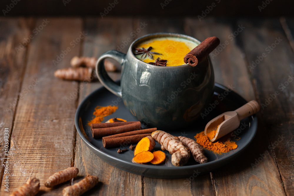 Golden milk in grey mug. Turmeric latte made with curcuma, cinnamon, anise. Healthy hot winter drink, natural, organic beverage. Close up, front view. Wooden rustic background. Raw roots as decor - obrazy, fototapety, plakaty 