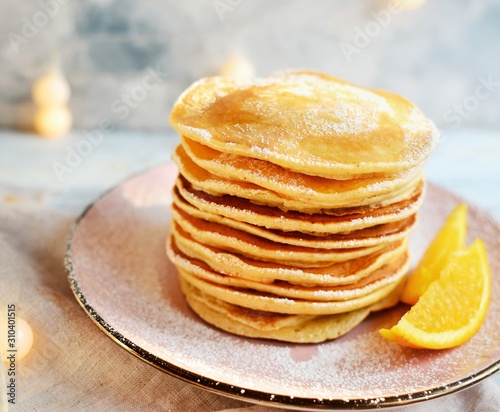 Pancakes with icing sugar and orange. Pancakes in a beautiful pink plate on a light wooden background. Delicious sweet food. Holiday pancakes and Christmas garland on the background.