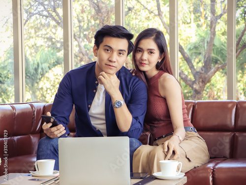 Happy Asian couple sitting on couch in living room with computer laptop and coffee cup on table smiling and looing to camera.