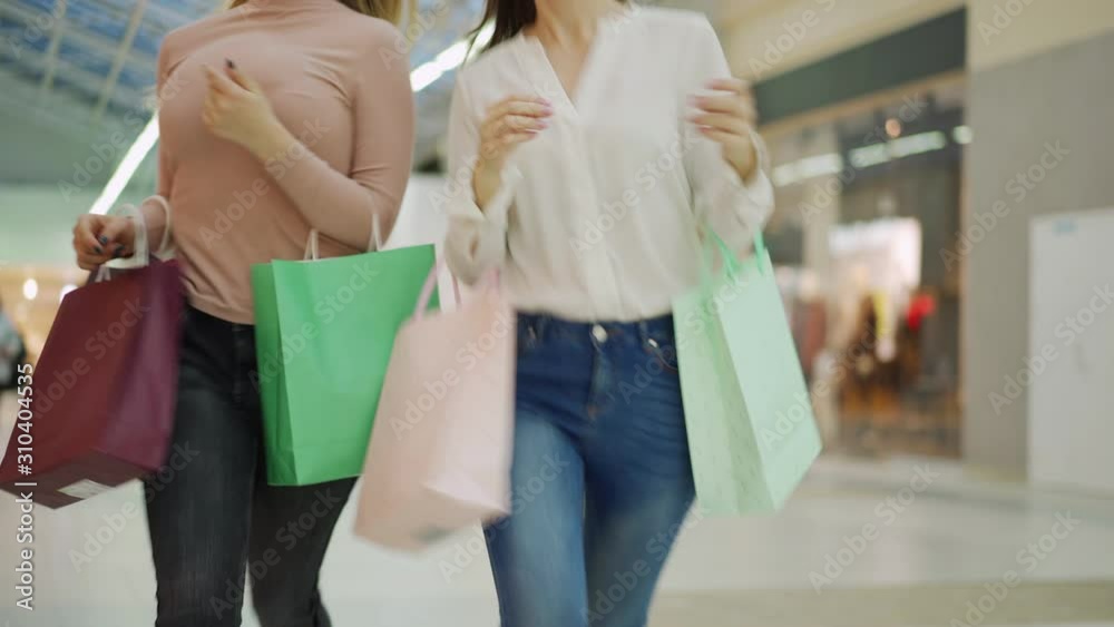 Medium shot of two unrecognizable faceless shopaholic women with shopping bags running along mall hurrying to make purchases on sale day Medium shot of two unrecognizable faceless shopaholic women with shopping bags running along mall hurrying to make purchases on sale day