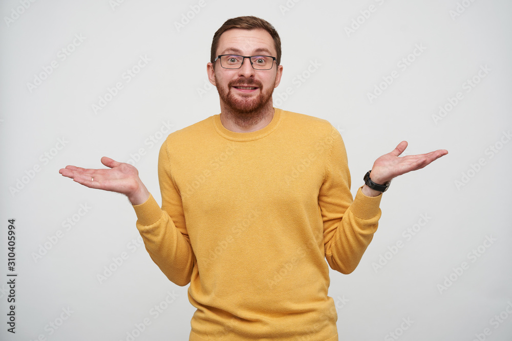 Studio photo of young handsome brunette bearded male shrugging with raised palms and smiling slightly at camera, wearing mustard sweater and wristwatch over white background Studio photo of young handsome brunette bearded male shrugging with raised palms and smiling slightly at camera, wearing mustard sweater and wristwatch over white background
