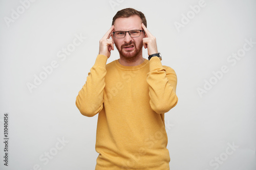 Displeased young brown haired man with beard wearing eyewear and mustard sweater while standing over white background, holding index fingers on his temples and frowning face © timtimphoto