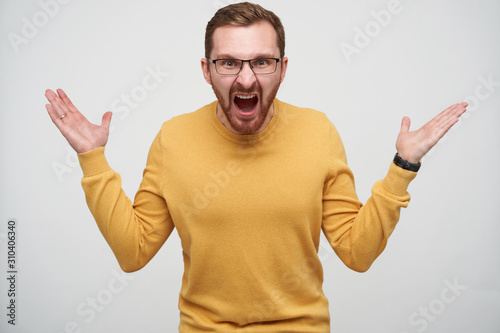 Studio photo of angry young brown haired bearded man in glasses raising emotionally palms up and shouting heatedly at camera, standing against white background