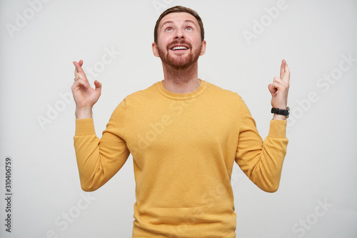 Cheerful young pretty brown haired bearded male with short haircut raising hands with crossed fingers and looking happily upwards, wearing mustard sweater over white background