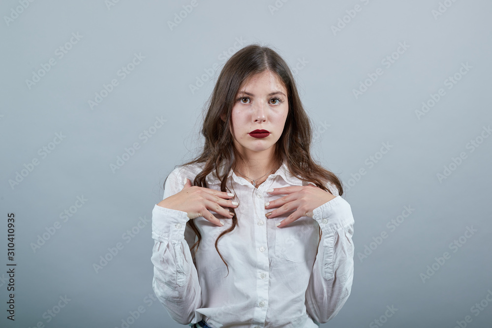 Serious young woman in fashion white shirt isolated on gray background in studio holding hans on chest, looking disappointed. People sincere emotions, lifestyle concept. Serious young woman in fashion white shirt isolated on gray background in studio holding hans on chest, looking disappointed. People sincere emotions, lifestyle concept.