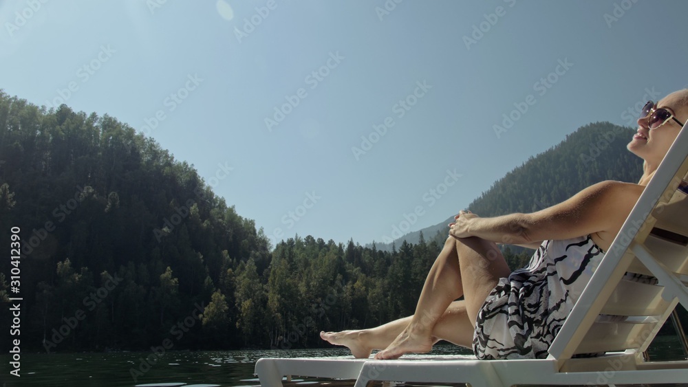 Woman lie on a sunbed in sunglasses and a boho silk shawl. Girl rest on a flood wood underwater pier. The pavement is covered with water in the lake. In the background are mountain and a forest.