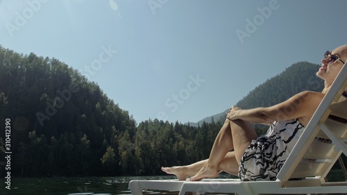 Woman lie on a sunbed in sunglasses and a boho silk shawl. Girl rest on a flood wood underwater pier. The pavement is covered with water in the lake. In the background are mountain and a forest.