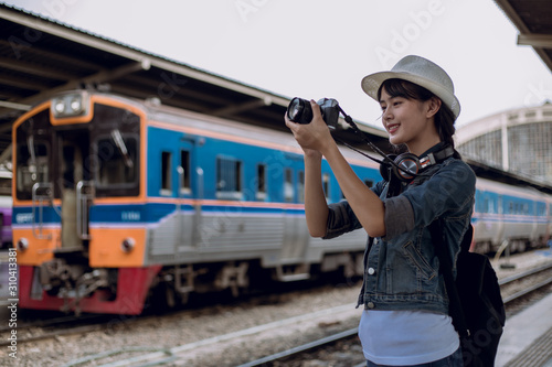 Side view of young woman smile, tourist girl with backpack taking photo of train station landscape view and smile happily. at the railway station, Backpacker concept, vintage train,blurred background.
