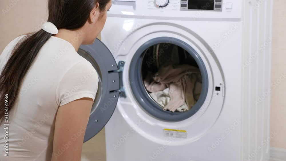long haired woman in t-shirt takes clean laundry of modern washing machine near beige wall slow motion closeup long haired woman in t-shirt takes clean laundry of modern washing machine near beige wall slow motion closeup