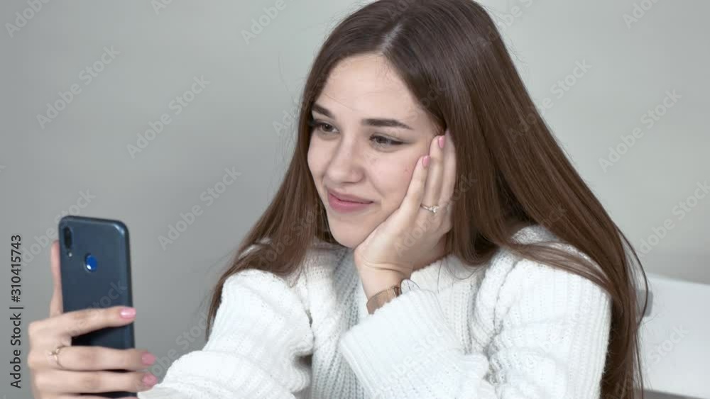 A young woman blogger communicates with her subscribers through a smartphone that she holds in her hand. She has long hair and a white sweater. She smiles. close-up A young woman blogger communicates with her subscribers through a smartphone that she holds in her hand. She has long hair and a white sweater. She smiles. close-up
