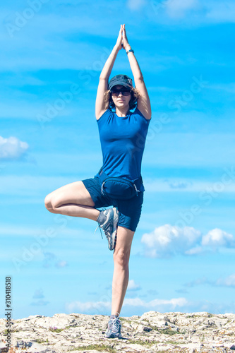 A girl in a tree pose from yoga exercises, standing on stones against a cloudy sky.