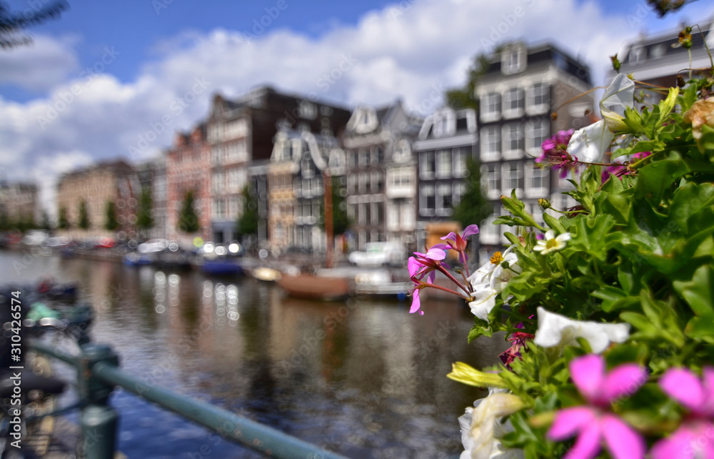 Fototapeta premium Amsterdam,Holland,August 2019. Enchanting view of a canals of the historic center. The typical houses overlook the water, boats moored along the canal. Planters on railings and bikes. Sunny day.