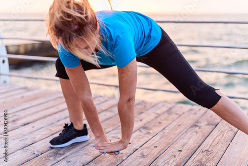 Woman with long blonde hair in blue shirt and black leggins doing stretching exercise deep side lunge withforward bend on seashore at sunrise