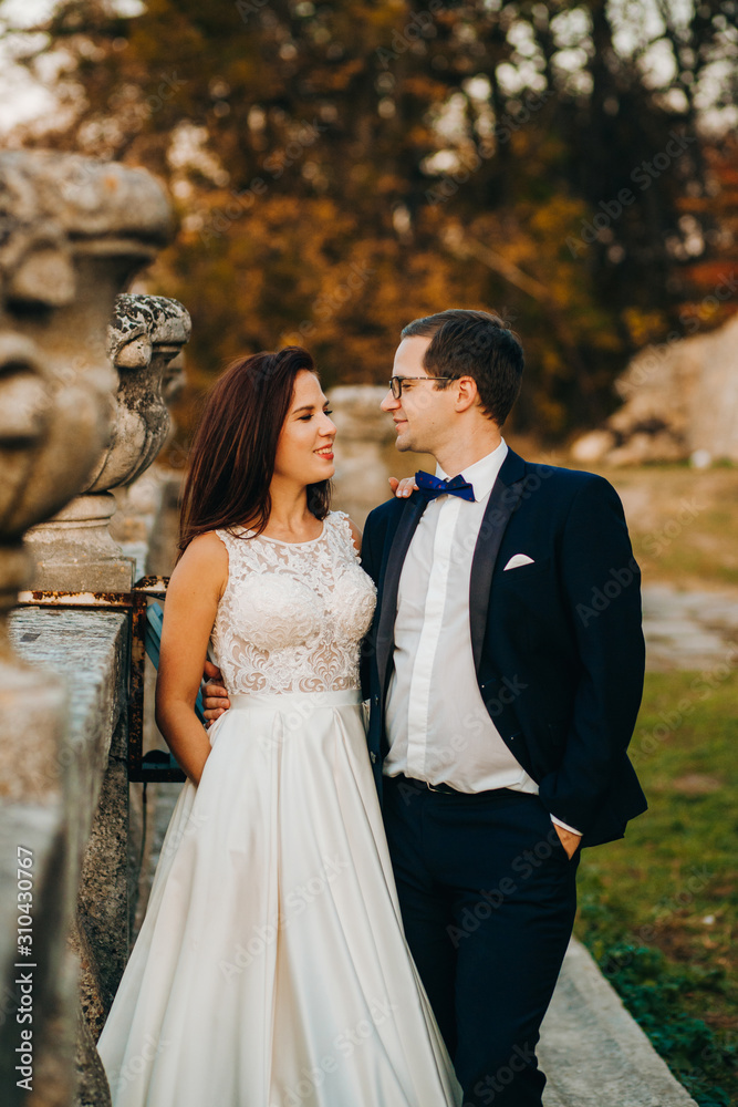 Sensual groom hugging newlywed bride from behind at old castle balcony with autumn trees background