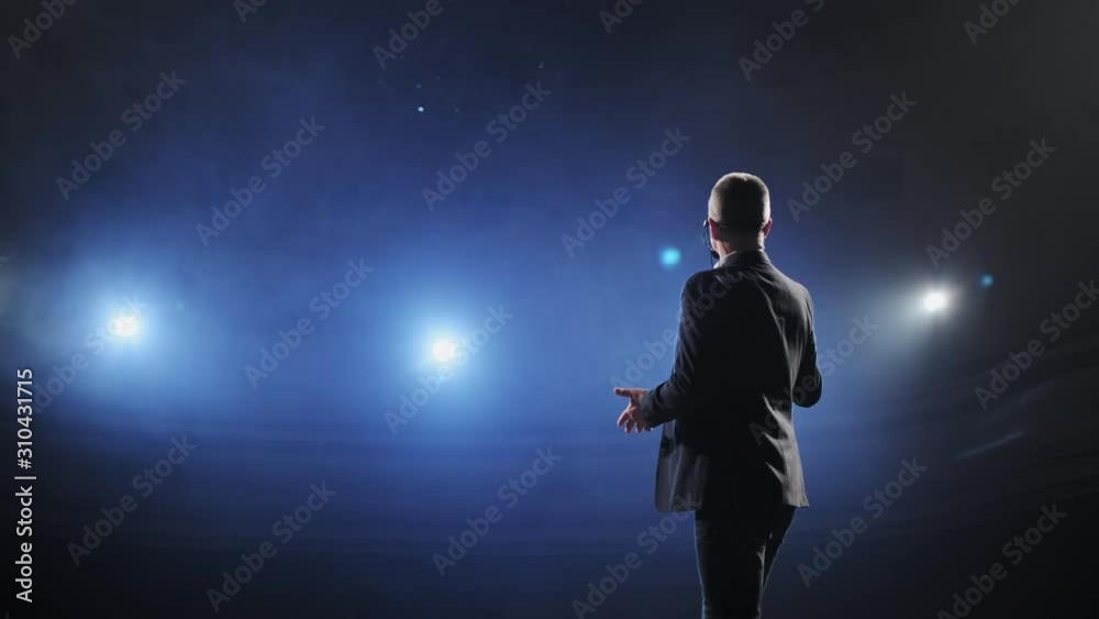 Showman. Middle Age Male entertainer, presenter or actor on stage. Arms to sides, smoke on background of spotlight. Rear view portrait of a male public speaker speaking at the microphone, pointing Showman. Middle Age Male entertainer, presenter or actor on stage. Arms to sides, smoke on background of spotlight. Rear view portrait of a male public speaker speaking at the microphone, pointing
