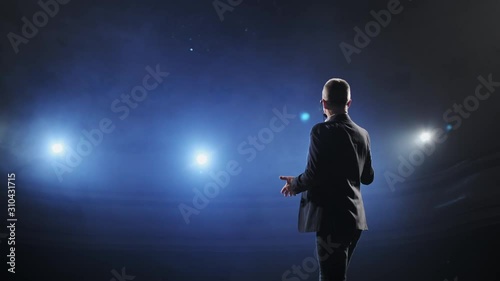 Showman. Middle Age Male entertainer, presenter or actor on stage. Arms to sides, smoke on background of spotlight. Rear view portrait of a male public speaker speaking at the microphone, pointing photo