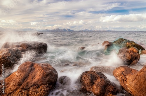 Mediterranean sea crashing onto rocks at Platja des Calo near Betlem and Colonia De Sant Pere with Cap De Formentor in the background, blue sky and white clouds, Mallorca, Spain.