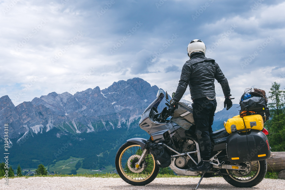 Naklejka premium Back view of stylish biker on adventure touring motorcycle in full equipment on dirt road, Look at distance on top of Dolomites mountains, travel concept, copy space. Cortina Ampezzo, Italy