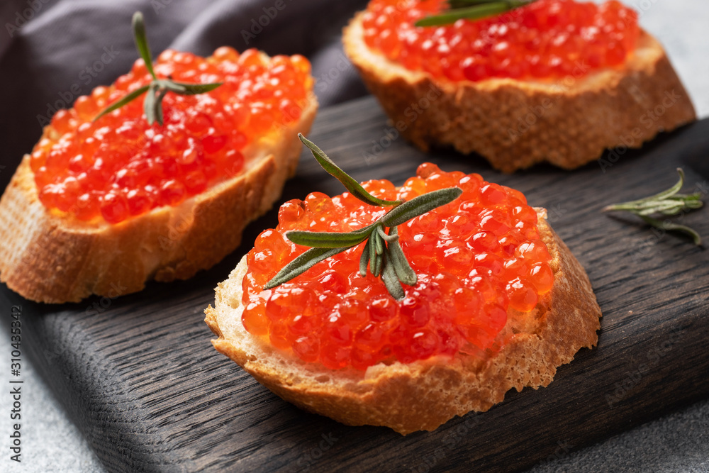 Sandwiches with red caviar on a white baguette with cottage cheese and rosemary. Dark wooden cutting Board. Grey concrete background. Close up. Sandwiches with red caviar on a white baguette with cottage cheese and rosemary. Dark wooden cutting Board. Grey concrete background. Close up.