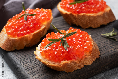 Sandwiches with red caviar on a white baguette with cottage cheese and rosemary. Dark wooden cutting Board. Grey concrete background. Close up.