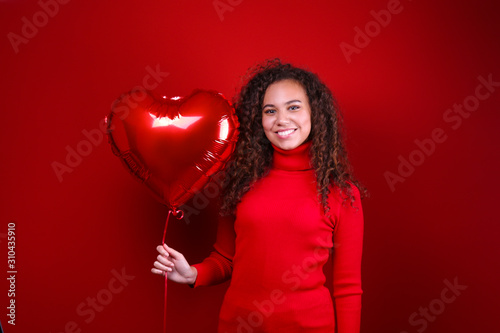 Studio portrait of young woman with dark skin and long curly hair wearing knitted turtle neck sweater over the festive red wall with heart shaped balloon. Close up, isolated background, copy space.