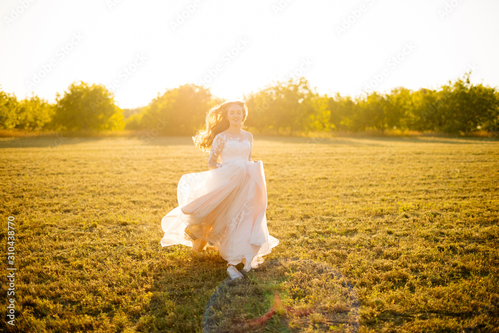 Young happy bride is running in front of the camera on the field at sunset.