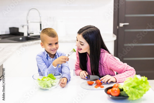Young mother with son boy cooking salad mom sliced vegetables food son give to mother tasting salad. Happy family cook food enjoyment lifestyle kitchen