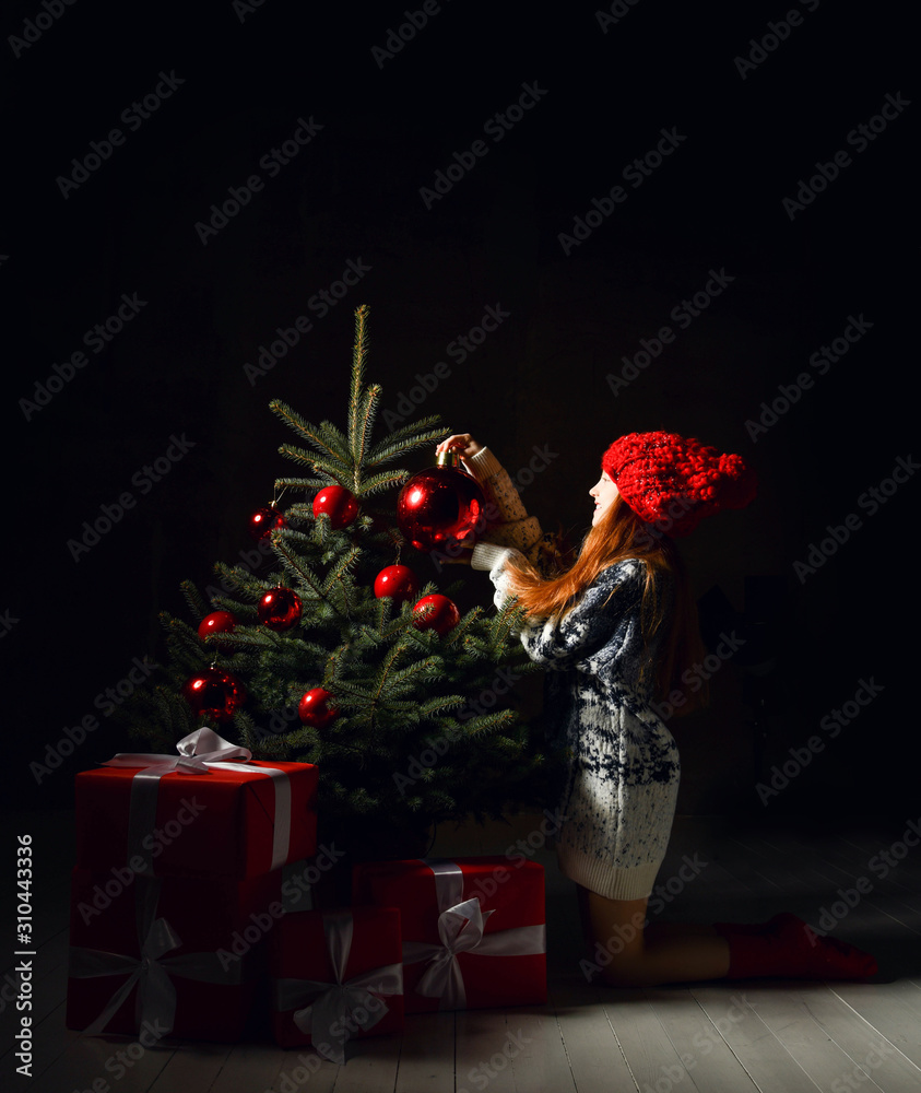 Young girl sitting celebrating decorates a christmas tree in red winter hat and presents gift boxes. New year Xmas 