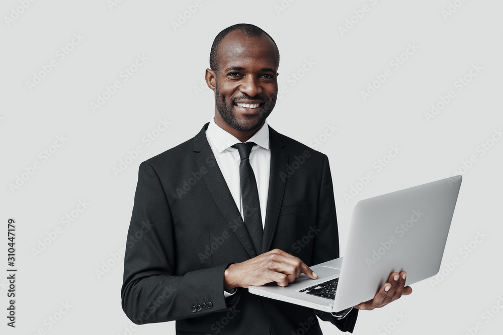 Confident young African man in formalwear working using computer while standing against grey background Confident young African man in formalwear working using computer while standing against grey background