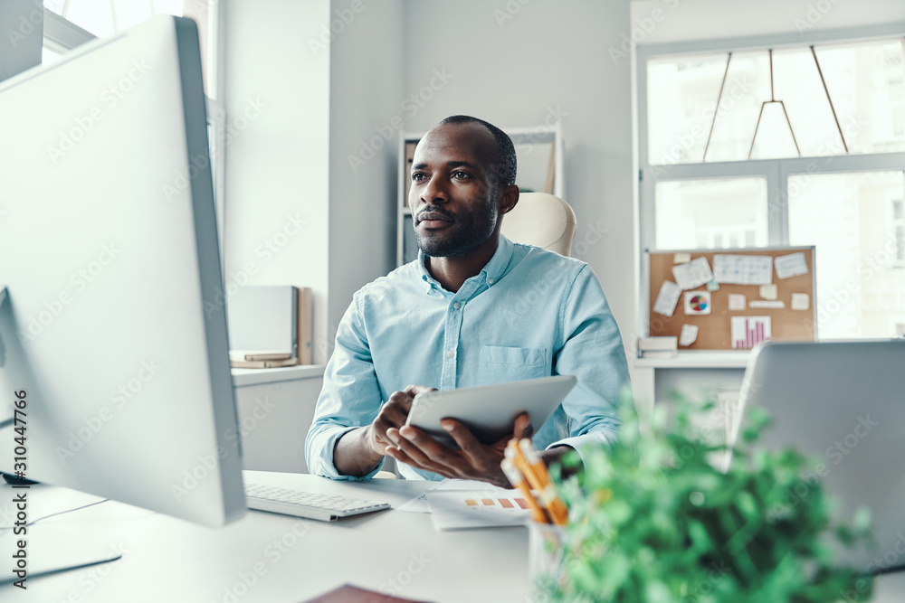 Thoughtful young African man in shirt using digital tablet while working in the office Thoughtful young African man in shirt using digital tablet while working in the office