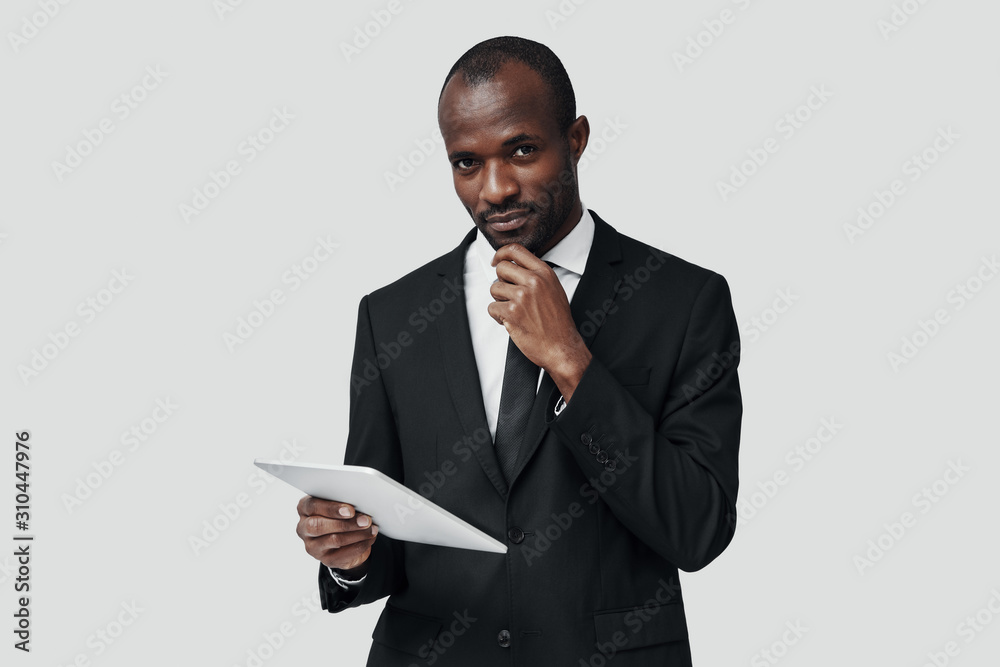 Successful young African man in formalwear working using digital tablet while standing against grey background Successful young African man in formalwear working using digital tablet while standing against grey background