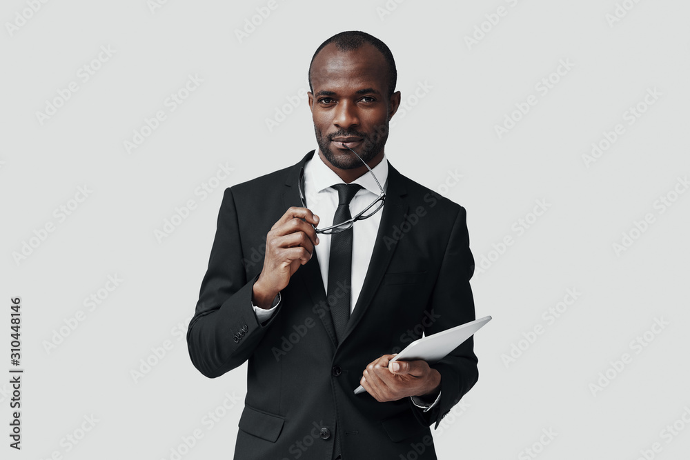 Charming young African man in formalwear working using digital tablet while standing against grey background Charming young African man in formalwear working using digital tablet while standing against grey background